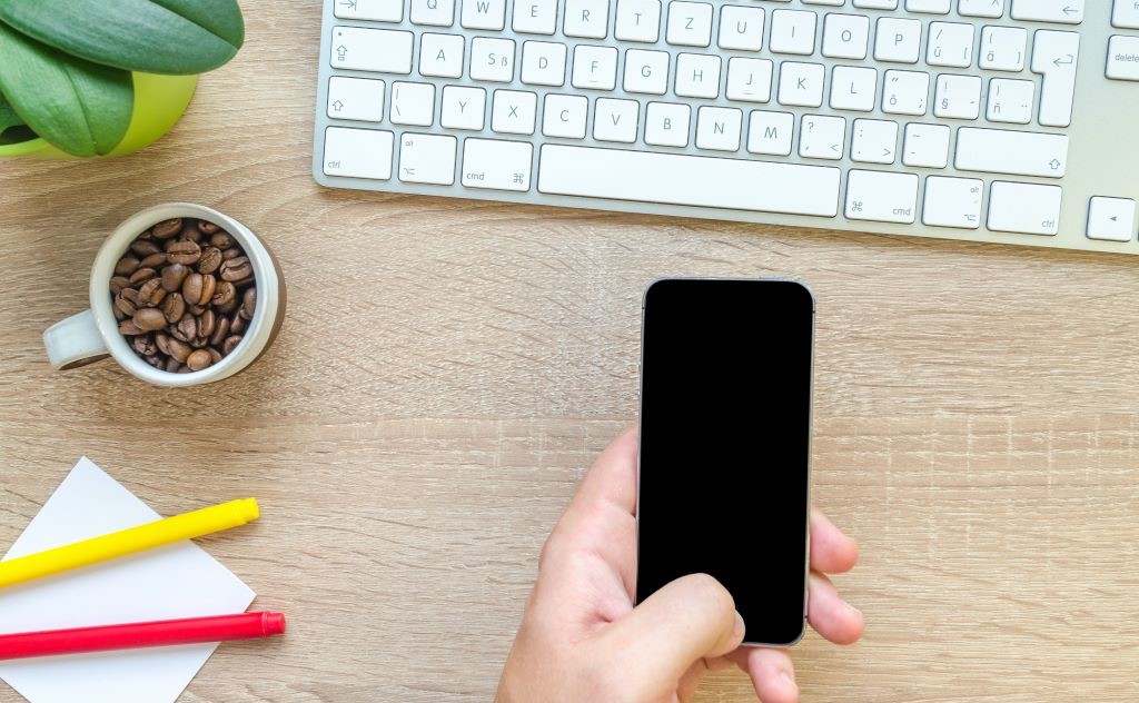 A desk setup, featuring a keyboard, some pens, a plant, a cup of coffee and a hand holding a mobile phone