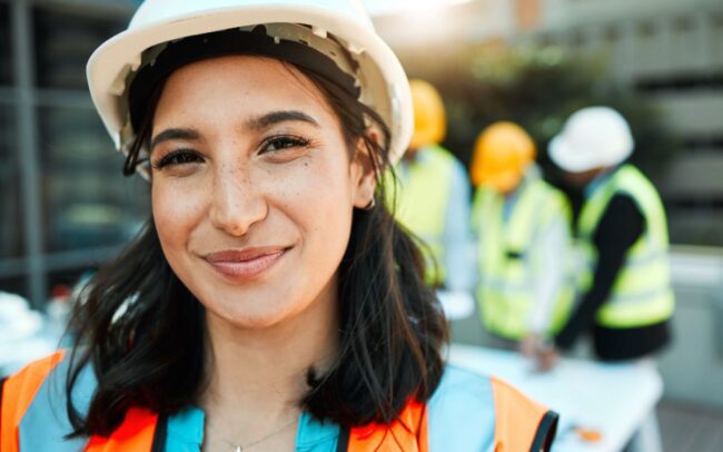 A construction worker, wearing a hard hat and hi-viz jacket smiles at the camera with three of her colleagues standing behind her.