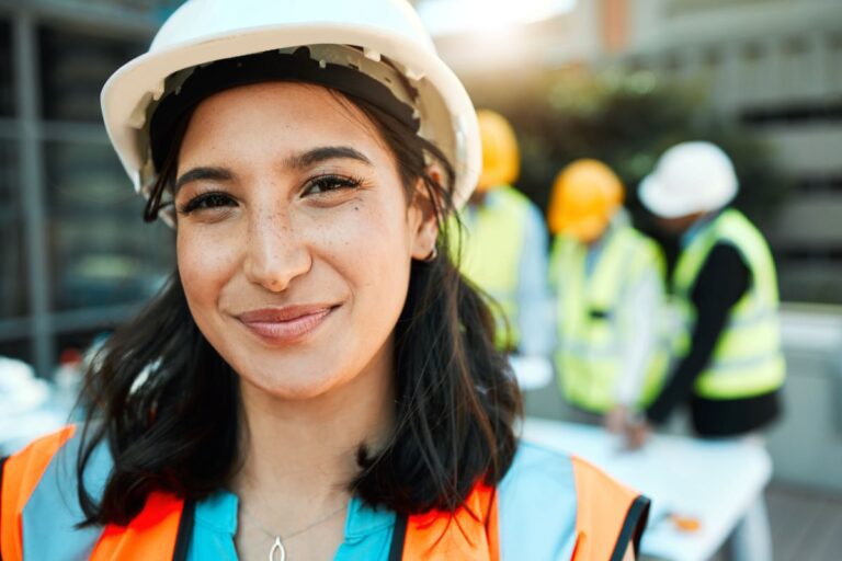 A construction worker, wearing a hard hat and hi-viz jacket smiles at the camera with three of her colleagues standing behind her.